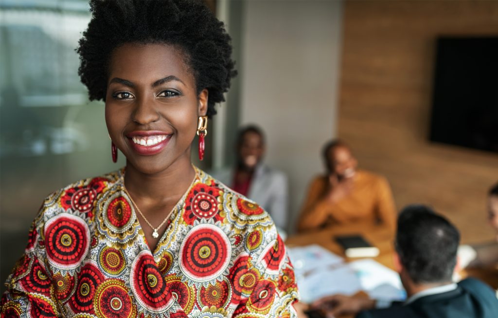 Lady all smiles in an office while her colleagues are seated behind her conversing