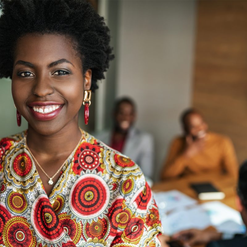 Lady all smiles in an office while her colleagues are seated behind her conversing