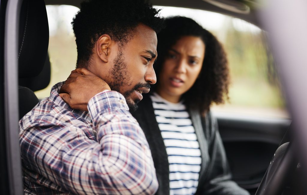 gent and lady in a car while the gent has his arm on the neck with frowny face as one in pain