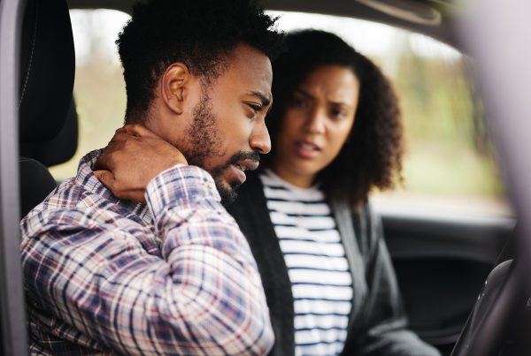 gent and lady in a car while the gent has his arm on the neck with frowny face as one in pain