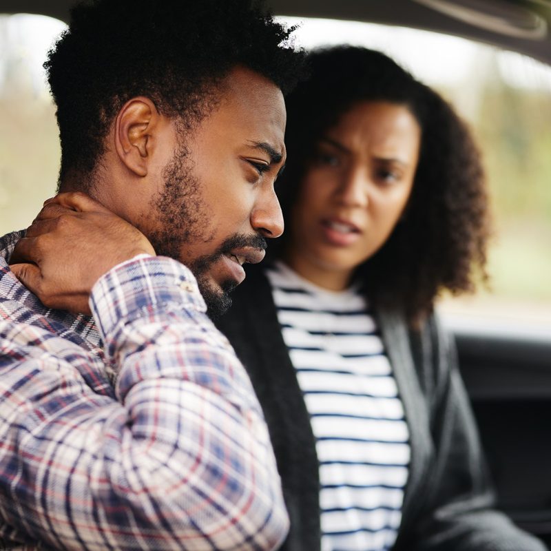 gent and lady in a car while the gent has his arm on the neck with frowny face as one in pain